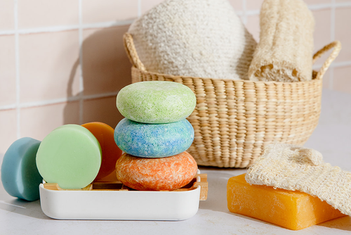 Colorful shampoo and conditioner bars stacked on an eco-friendly soap dish next to an agave soap saver bag resting on top of an orange soap bar