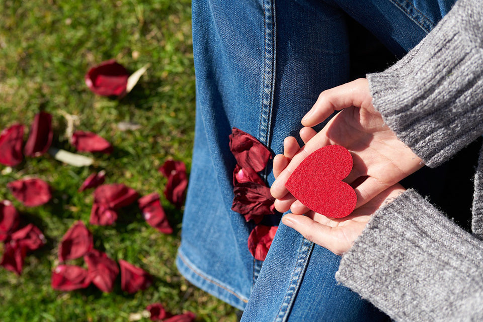 Person holding heart shaped cutout surrounded by rose petals on grass