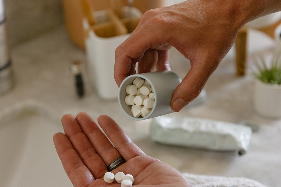 one hand pouring out small white toothpaste tablets from a light blue tin into another hand