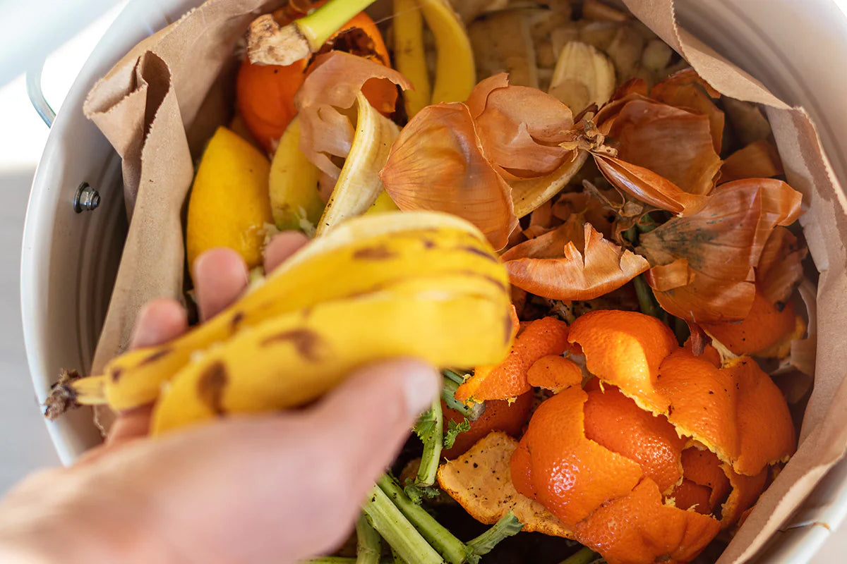 adding a banana peel to a large metal container of other vegetable and fruit scraps and peels