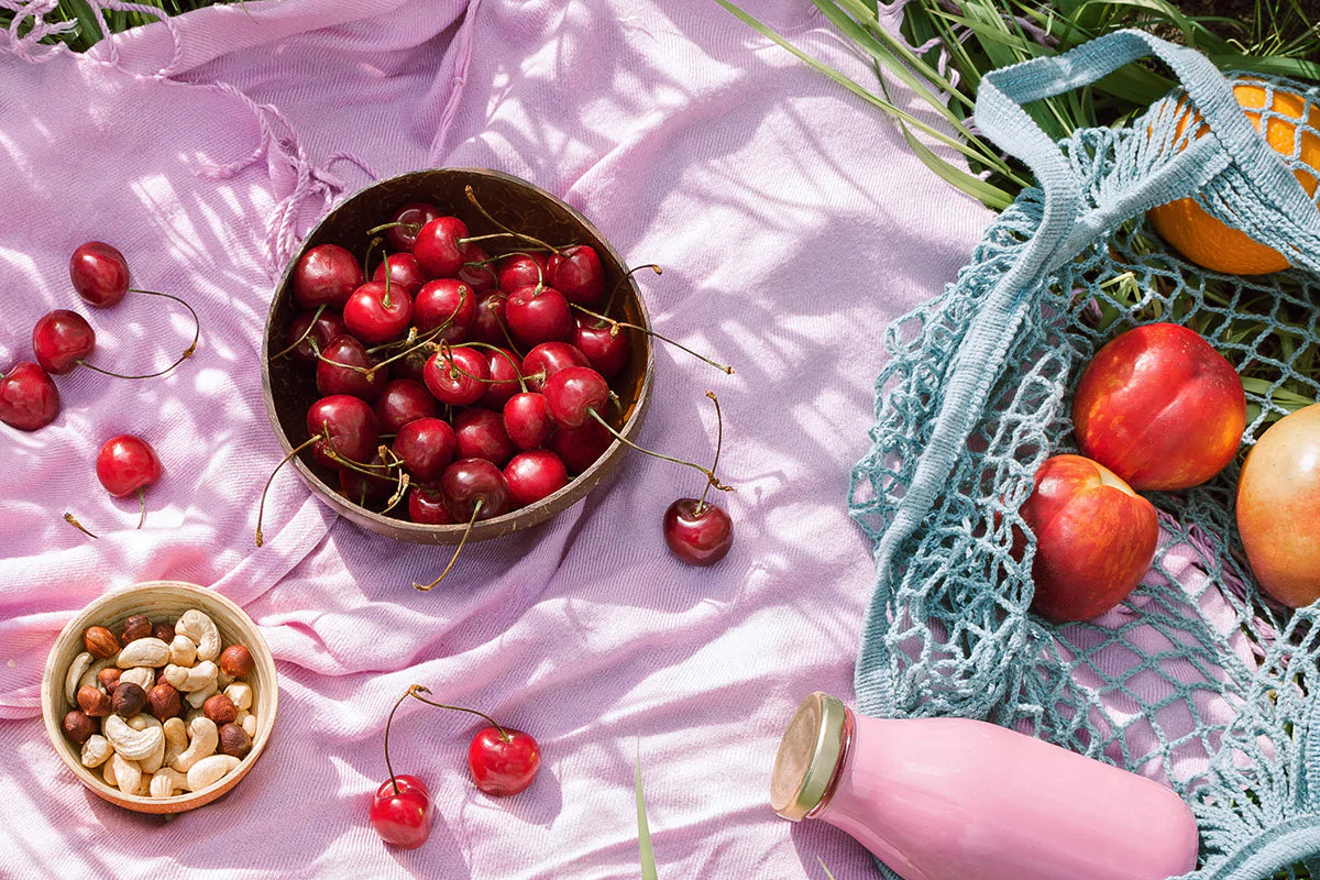 zero waste picnic setup on a pink scarf, with wooden bowls of cherries and mixed nuts, and a blue net bag of fresh fruit