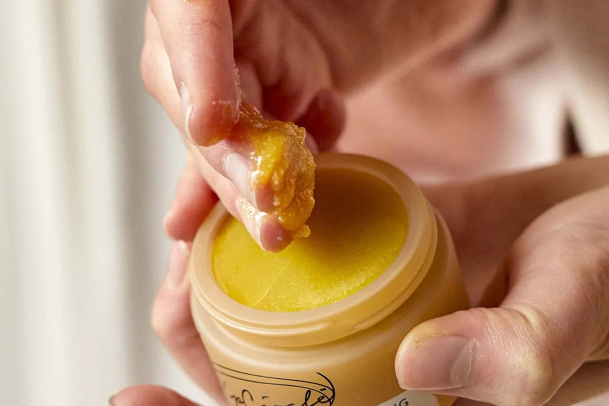 hand scooping product from a small jar of yellow-colored facial scrub